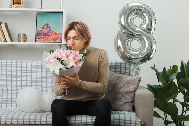 Erfreut mit geschlossenen Augen, gutaussehender Kerl am glücklichen Frauentag, der einen Blumenstrauß auf dem Sofa im Wohnzimmer hält