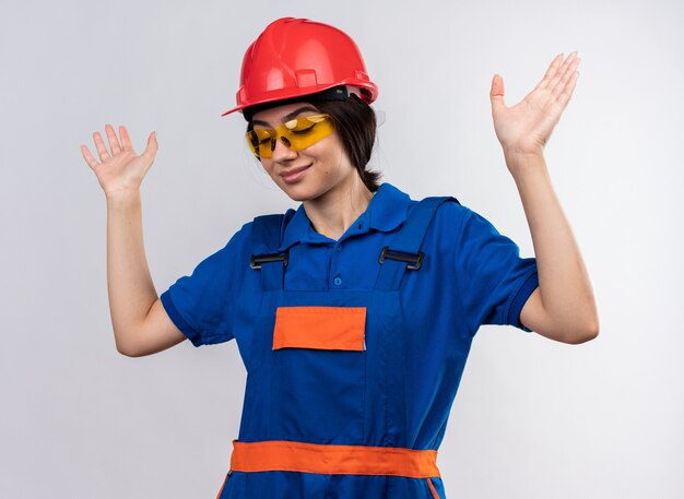 Erfreut mit geschlossenen Augen junge Baumeisterin in Uniform und Brille, die die Hände isoliert auf weißer Wand hebt
