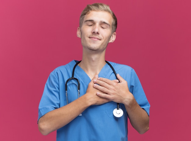 Erfreut mit geschlossenen Augen junger männlicher Arzt, der Arztuniform mit Stethoskop trägt, die Hände auf Herz lokalisiert auf rosa Wand setzen