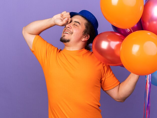 Erfreut mit geschlossenen Augen junger Mann mit Partyhut, der Luftballons hält und das Auge mit der Hand isoliert auf lila Wand abwischt