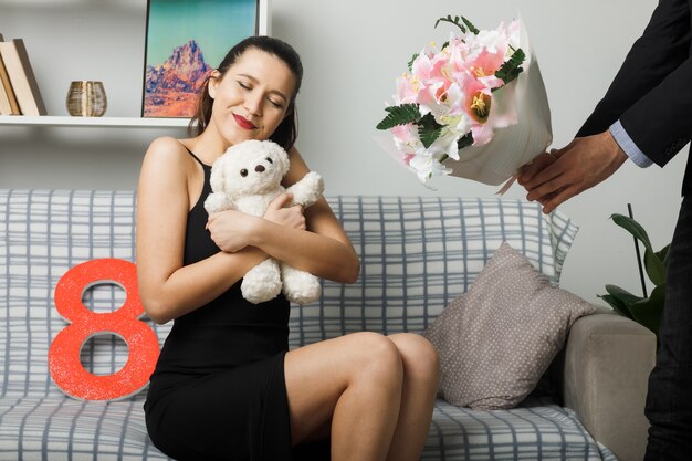 Erfreut mit geschlossenen Augen junges Mädchen am glücklichen Frauentag, der auf dem Sofa sitzt und Teddybär im Wohnzimmer hält