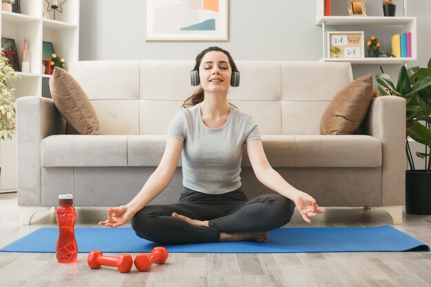 Erfreut mit geschlossenen Augen junges Mädchen mit Kopfhörern trainiert auf Yogamatte vor dem Sofa im Wohnzimmer