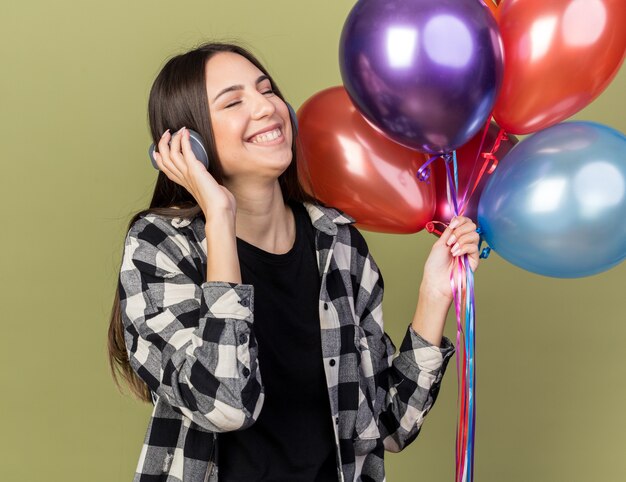 Erfreut mit geschlossenen Augen junges schönes Mädchen mit Kopfhörern mit Luftballons isoliert auf olivgrüner Wand