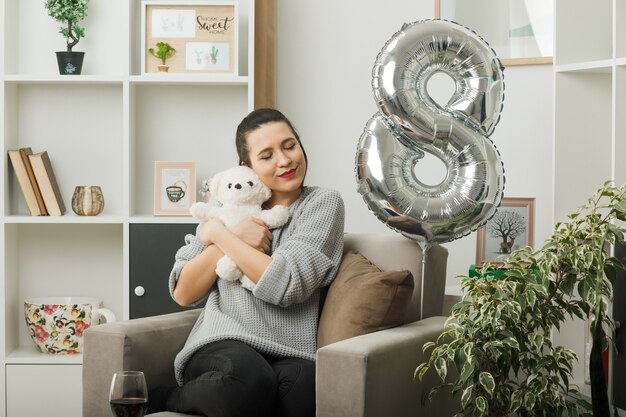 Erfreut mit geschlossenen Augen schöne Frau am glücklichen Frauentag mit Teddybär sitzend auf Sessel im Wohnzimmer