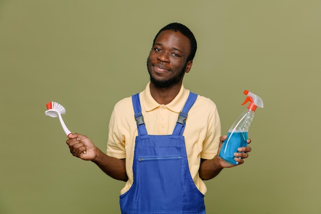 Erfreut, Reinigungsmittel mit Reinigungsbürste zu halten Junge afroamerikanische Reinigungskraft in Uniform mit Handschuhen isoliert auf grünem Hintergrund