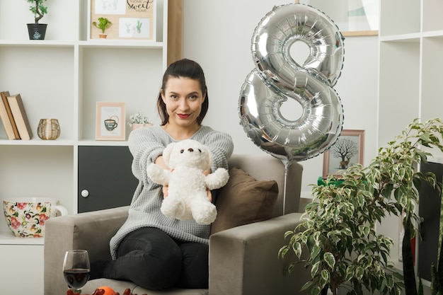 Erfreut schöne Frau am glücklichen Frauentag, der den Teddybären in die Kamera hält, der auf einem Sessel im Wohnzimmer sitzt