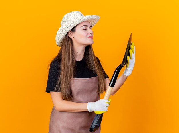 Erfreut schönes Gärtnermädchen, das Uniform und Gartenhut mit Handschuhen hält, die Spaten lokalisiert auf orange Hintergrund halten und betrachten