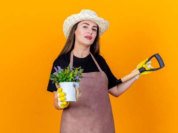 Erfreut schönes Gärtnermädchen, das Uniform und Gartenhut mit Handschuhen trägt, die Spaten auf Schulter setzen und Blume im Blumentopf halten, der auf orange Hintergrund lokalisiert wird