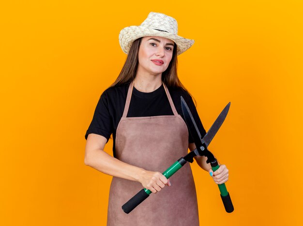 Erfreut schönes Gärtnermädchen, das Uniform und Gartenhut trägt, die Clipper lokalisiert auf Orange halten