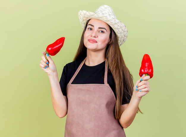 Erfreut schönes Gärtnermädchen in der Uniform, die Gartenhut hält, der Paprika lokalisiert auf olivgrünem Hintergrund hält