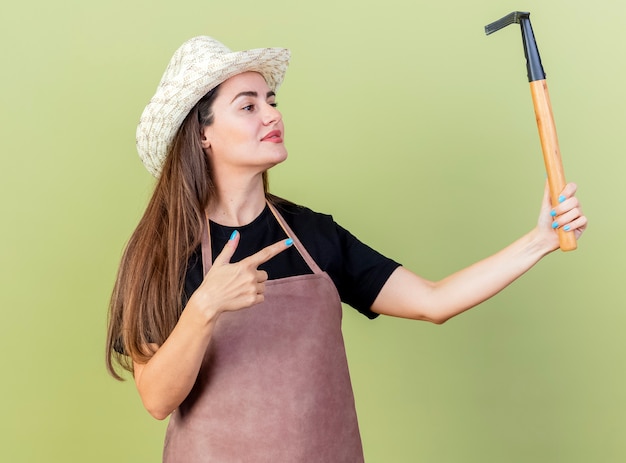 Erfreut schönes Gärtnermädchen in der Uniform, die Gartenhut hält und Punkte auf Rechen lokalisiert auf Olivgrün trägt