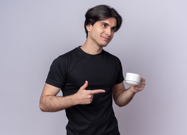 Erfreut, seitlichen jungen gutaussehenden Mann zu betrachten, der schwarze T-Shirt hält und Punkte auf Tasse Kaffee lokalisiert auf weißer Wand zeigt