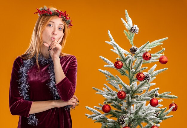 Erfreut spitzen Lippen junges schönes Mädchen, das nahe Weihnachtsbaum steht, das rotes Kleid und Kranz mit Girlande am Hals trägt Finger auf Wange lokalisiert auf orange Hintergrund