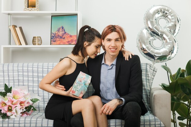 Erfreut umarmten sich junge Paare am glücklichen Frauentag, der das Geschenk auf dem Sofa im Wohnzimmer hielt