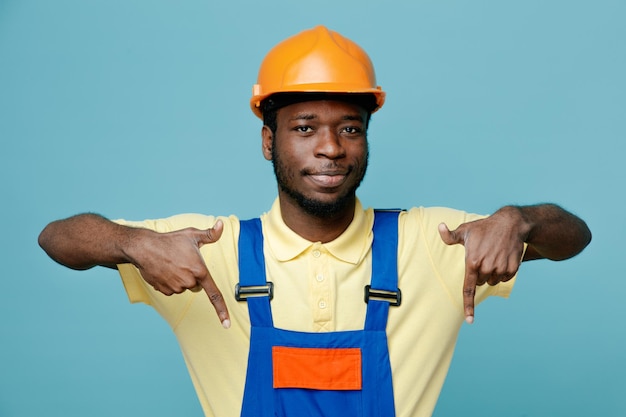 Erfreut zeigt auf den jungen afroamerikanischen Baumeister in Uniform, der auf blauem Hintergrund isoliert ist