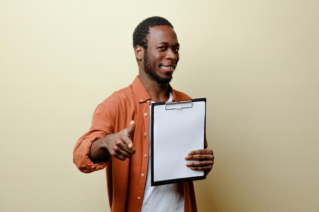 Erfreut zeigt auf die Kamera junger Afroamerikaner, der die Zwischenablage isoliert auf weißem Hintergrund hält