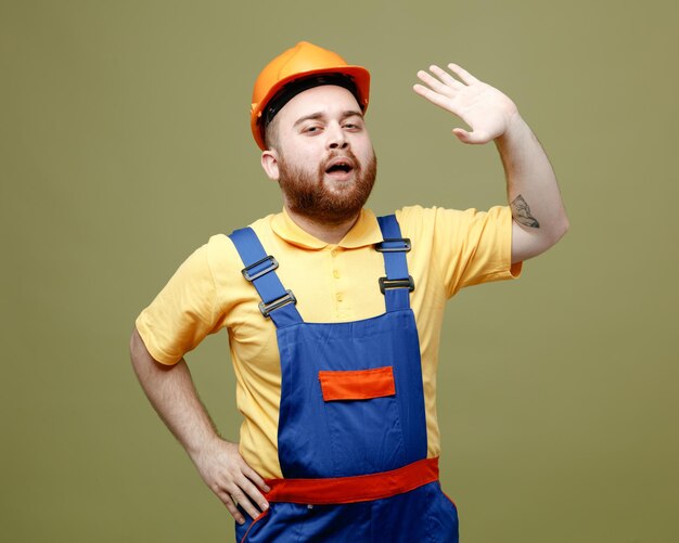 Erfreut zeigt hallo Geste junger Baumeister Mann in Uniform isoliert auf grünem Hintergrund
