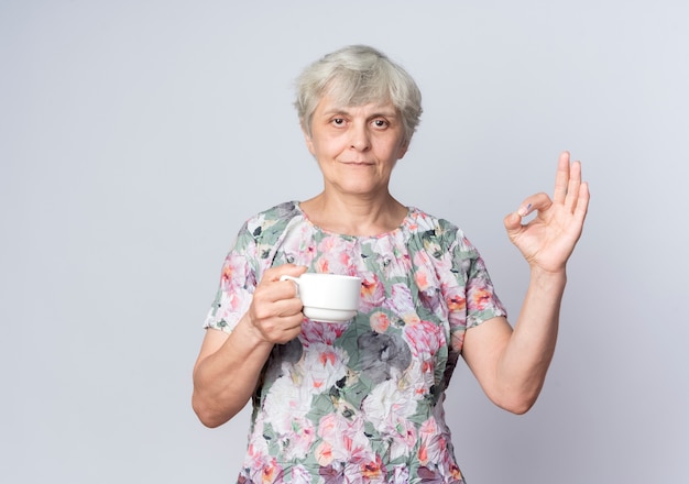 Erfreute ältere Frau hält Tasse und gestikuliert ok Handzeichen lokalisiert auf weißer Wand