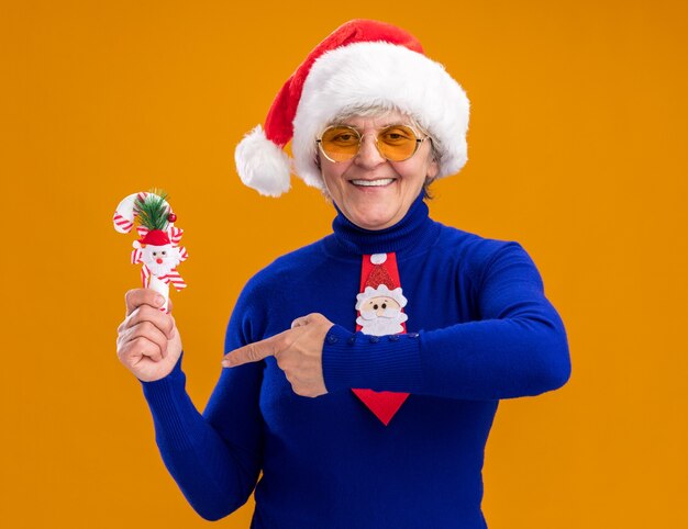 Erfreute ältere Frau in Sonnenbrille mit Santa Hut und Santa Tie hält und zeigt auf Zuckerstange lokalisiert auf orange Hintergrund mit Kopienraum