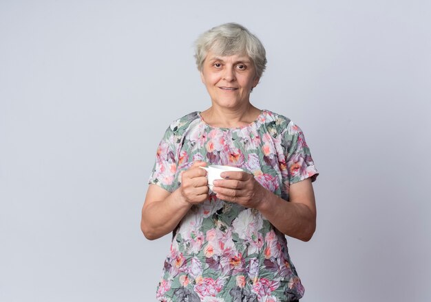 Erfreute ältere Frau steht hält Tasse lokalisiert auf weißer Wand