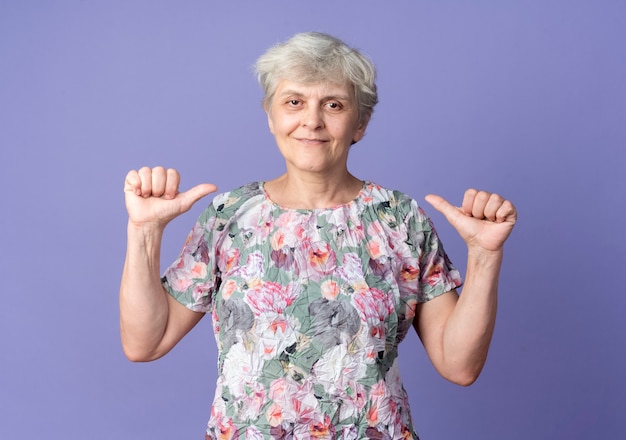 Erfreute ältere Frau zeigt auf sich selbst mit zwei Händen, die auf lila Wand isoliert sind