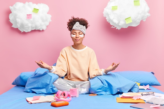 Erfreute Afroamerikanerin meditiert im Bett, fühlt sich entspannt, trägt Nachtwäsche, schließt die Augen und praktiziert Yoga, nachdem sie Kaffee getrunken hat