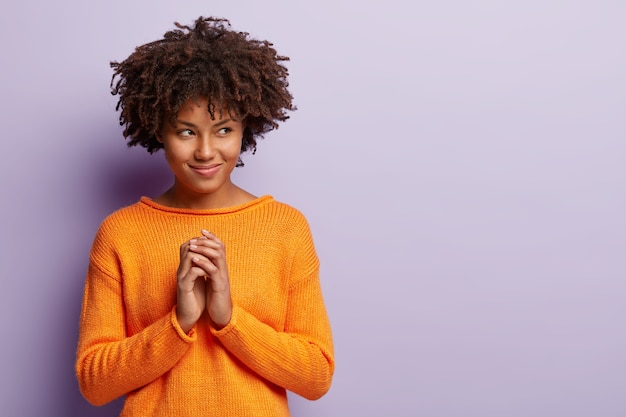 Erfreute afroamerikanische Frau macht Plan, hat faszinierenden Blick auf freien Raum beiseite, hält Handflächen zusammen, schaut weg, trägt orangefarbenen Pullover, isoliert über lila Wand. Menschen, Absichtskonzept.