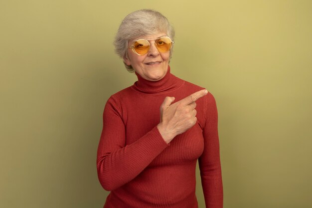 Erfreute alte Frau mit rotem Rollkragenpullover und Sonnenbrille, die auf die Seite zeigt, isoliert auf olivgrüner Wand mit Kopierraum