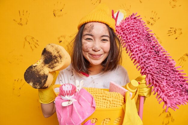Erfreute asiatische Frau hat Freude am Reinigungsprozess hält schmutzigen Schwamm und Mopp in der Nähe eines Korbes voller Wäsche isoliert über gelber Wand