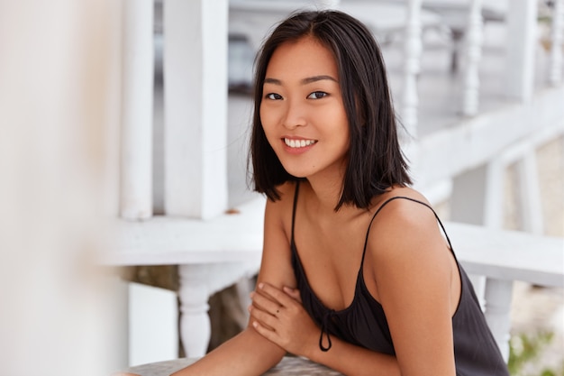 Erfreute asiatische Frau mit breitem Lächeln hat Frisur geschaukelt, lässig gekleidet, sitzt am Kaffeetisch, genießt Freizeit. Schöne japanische Frau ruht allein im Restaurant