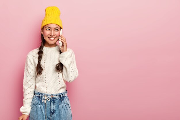 Erfreute asiatische Frau spricht über Smartphone, hat angenehme Gespräche, ruft Freund an, trägt warmweißen Pullover und Jeans, schaut mit einem Lächeln zur Seite, posiert über rosa Wand