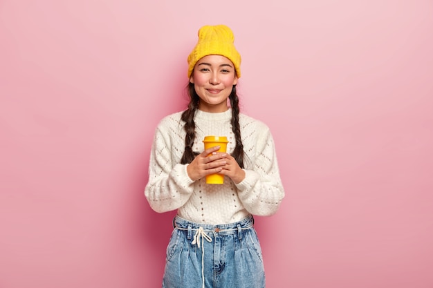 Erfreute attraktive Frau mit Zöpfen, gut gekleidet, trinkt gerne Kaffee aus der Tasse zum Mitnehmen, hat fröhlichen Ausdruck, posiert über rosa Wand