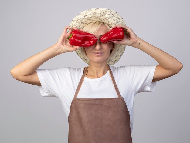 Erfreute blonde Gärtnerin mittleren Alters in Uniform mit Hut, die Paprika vor den Augen isoliert auf weißer Wand hält