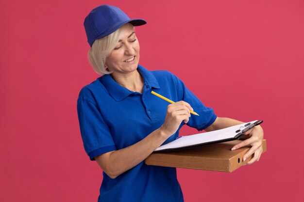 Erfreute blonde Lieferfrau mittleren Alters in blauer Uniform und Mütze mit Bleistift-Zwischenablage-Pizza-Paket mit Blick auf die Zwischenablage