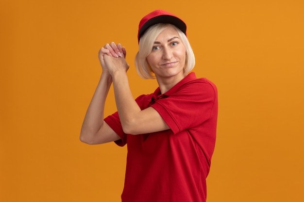 Erfreute blonde Lieferfrau mittleren Alters in roter Uniform und Mütze mit Blick auf die Vorderseite mit gewinnender Geste einzeln auf oranger Wand mit Kopierraum