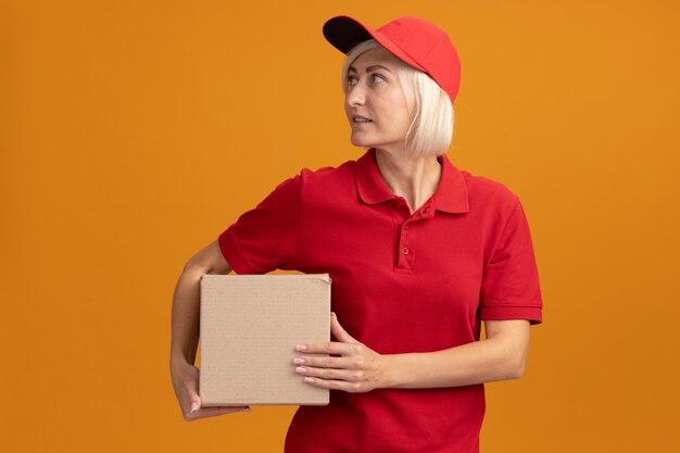 Erfreute blonde Lieferfrau mittleren Alters in roter Uniform und Mütze mit Karton mit Blick auf die Seite isoliert auf oranger Wand
