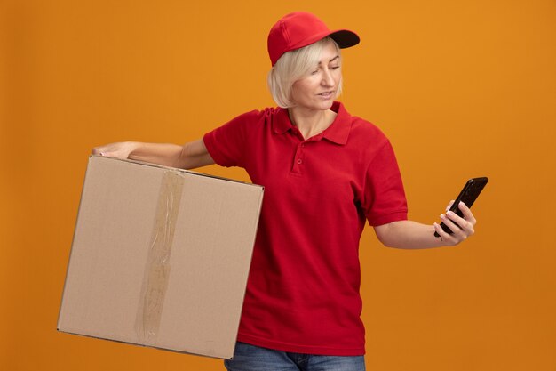 Erfreute blonde lieferfrau mittleren alters in roter uniform und mütze mit karton und handy, die auf das telefon schaut
