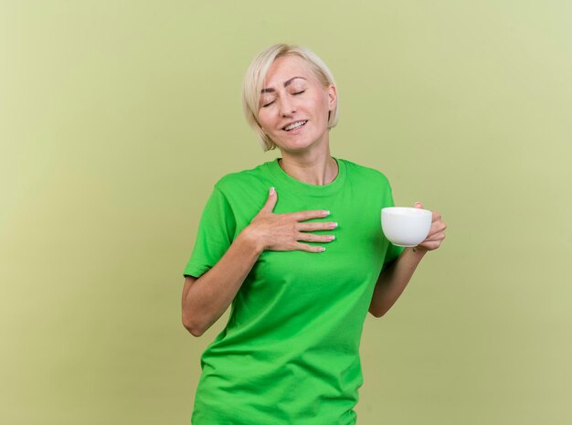 Erfreute blonde slawische Frau mittleren Alters, die eine Tasse Tee hält Hand auf Brust mit geschlossenen Augen lokalisiert auf olivgrüner Wand mit Kopienraum