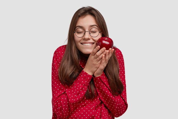 Erfreute Brünette Frau fühlt Vergnügen, hält leckeren Apfel, ist in Hochstimmung, trägt rote Kleidung