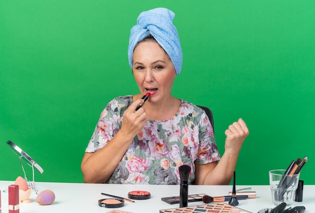 Erfreute erwachsene kaukasische Frau mit eingewickeltem Haar in Handtuch, die am Tisch mit Make-up-Tools sitzt und Lippenstift aufträgt