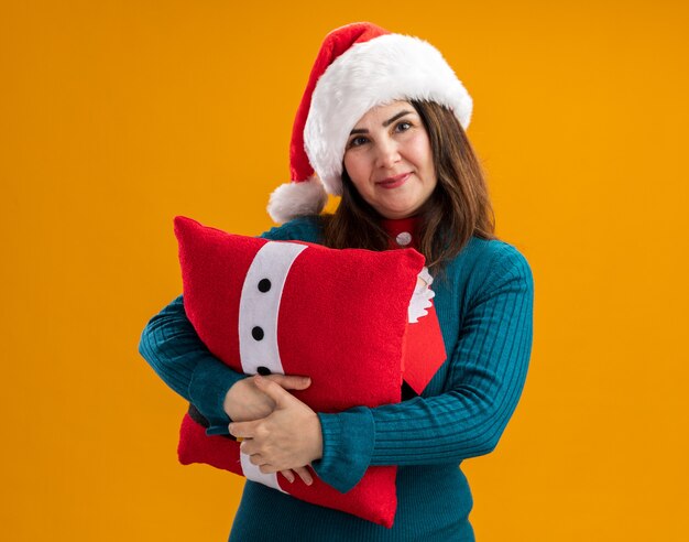 Erfreute erwachsene kaukasische Frau mit Weihnachtsmütze und Weihnachtsmann-Krawatte umarmt dekoriertes Kissen isoliert auf oranger Wand mit Kopierraum