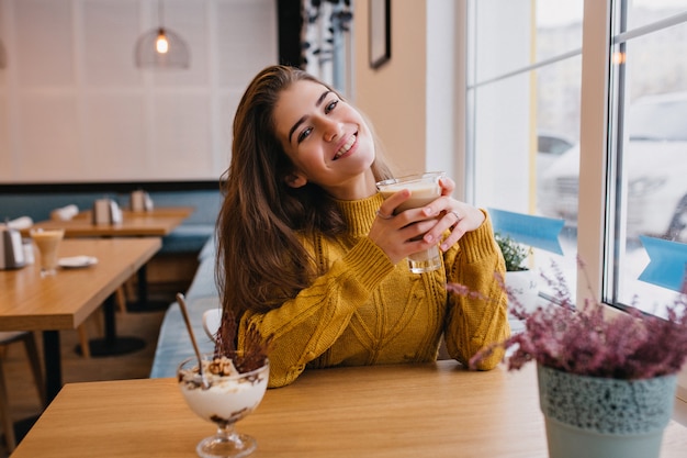 Erfreute Frau mit dunklem Haar, das im Winter mit Tasse Kaffee im gemütlichen Café kühlt. Innenporträt der erstaunlichen Dame in der gestrickten gelben Strickjacke, die im Restaurant ruht und Eis genießt.