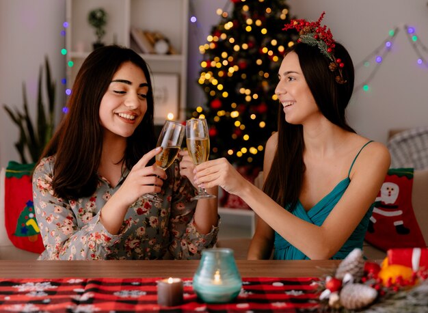 erfreute hübsche junge Mädchen klirren Gläser Champagner, die am Tisch sitzen und die Weihnachtszeit zu Hause genießen