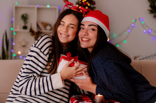 erfreute hübsche junge Mädchen mit Rentierbrille und Weihnachtsmütze halten Weihnachtsgeschenkbox sitzen auf Sesseln und genießen Weihnachtszeit zu Hause