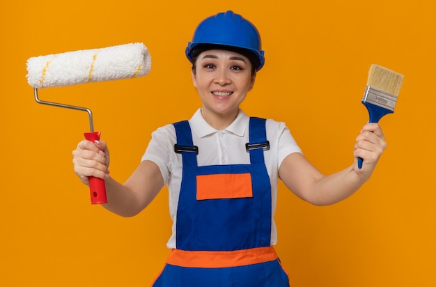 Erfreute junge asiatische Baumeisterin mit blauem Schutzhelm mit Farbroller und Pinsel