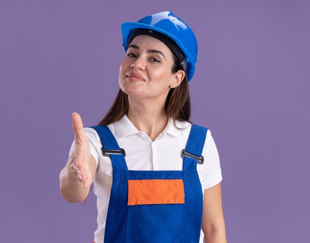 Erfreute junge Baumeisterin in Uniform, die Hand an der Kamera lokalisiert auf lila Wand heraushält