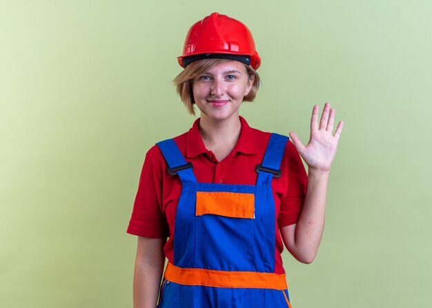 erfreute junge Baumeisterin in Uniform mit Hallo-Geste isoliert auf olivgrüner Wand