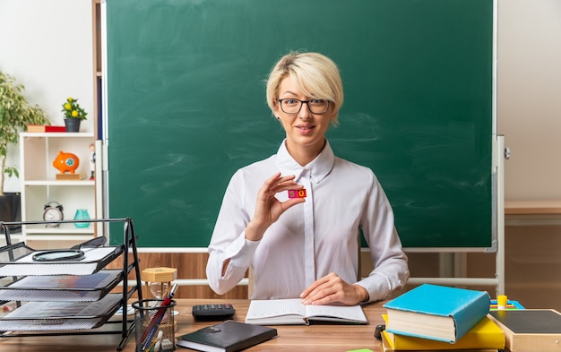 Erfreute junge blonde Lehrerin mit Brille, die am Schreibtisch mit Schulwerkzeugen im Klassenzimmer sitzt und kleine quadratische Zahlen fünf und null zeigt