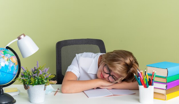 Erfreute junge blonde Studentin mit Brille am Schreibtisch sitzend mit Schulwerkzeugen, die mit geschlossenen Augen den Kopf auf dem Schreibtisch ruhen lassen