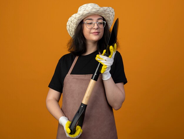 Erfreute junge brünette Gärtnerin in optischen Gläsern und in Uniform mit Gartenhut und Handschuhen, die Spaten lokalisiert auf orange Wand halten und betrachten